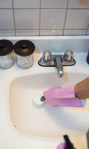 A person using a brush and pink gloves to clean a white bathroom sink, promoting hygiene and cleanliness.